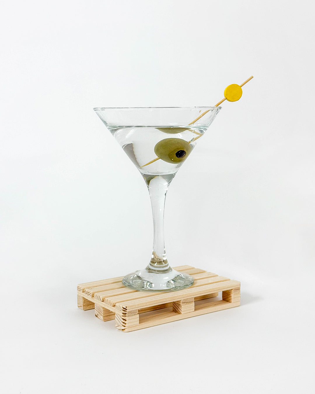 Martini kozarec z olivo postavljen na mini leseni paleti z belim ozadjem.