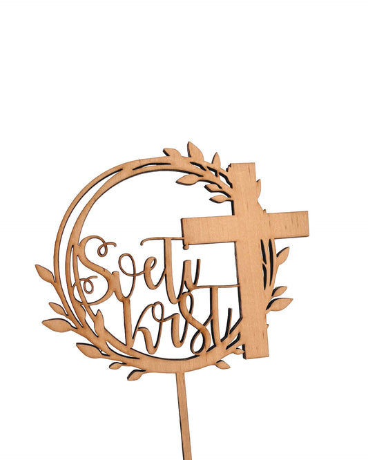 topper ali napis za torto sveti krst. Toper je na sredini bele visoke torte. Je iz temnega furnirja. Na toperju je desno velik križ, levo pa piše sveti krst. Vsaka beseda je v svoji vrstici. Toper za torto sveti krst.