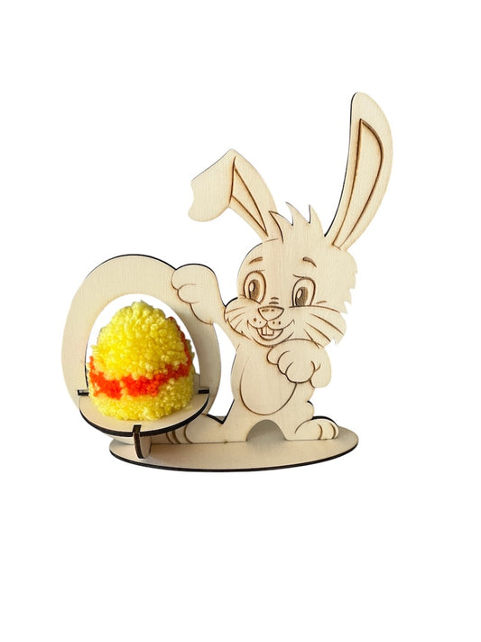 Leseni zajček s stojalom na levi strani, zajček se z eno roko drži stojala. V stojalu je rumeni pirh narejen iz pom pom. Rumeni pirh ima v sredini oranžno črto. Zajček in stojalo sta na ovalni leseni podlogi.